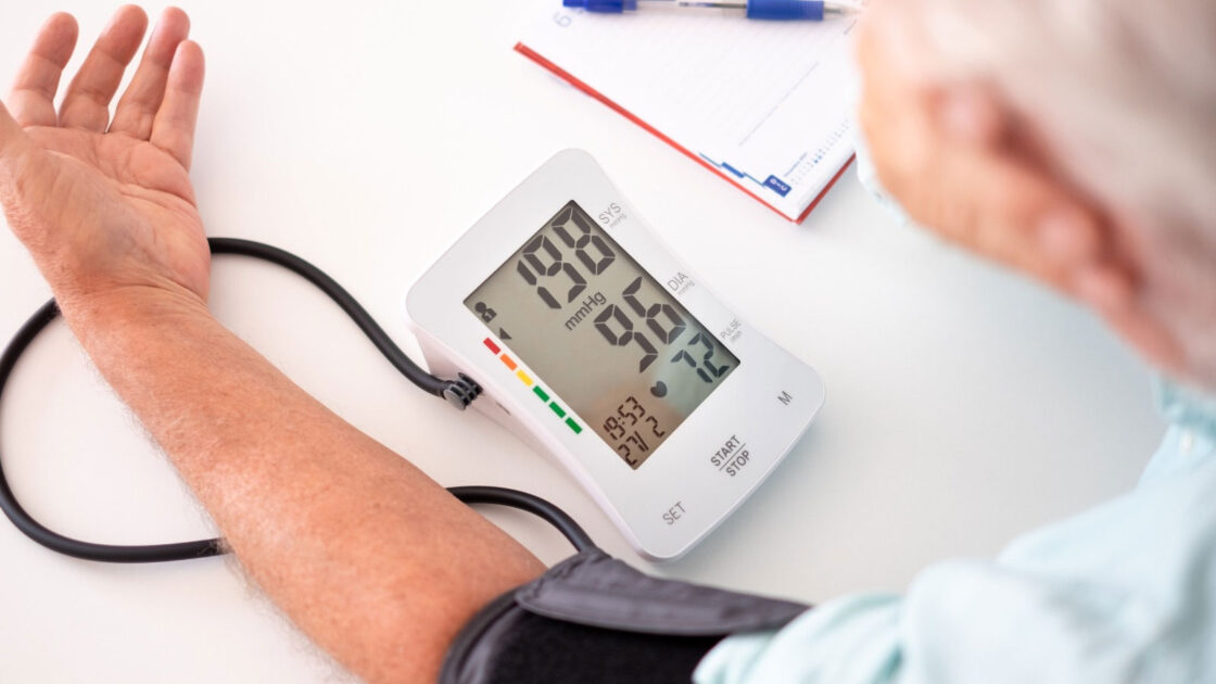 Digitālais asinsspiediena mērītājs uz vīrieša rokas uzrāda kritiski augstu asinsspiedienu 198 uz 96 mmHg, kas liecina par hipertensīvo krīzi.