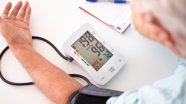 Digitālais asinsspiediena mērītājs uz vīrieša rokas uzrāda kritiski augstu asinsspiedienu 198 uz 96 mmHg, kas liecina par hipertensīvo krīzi.